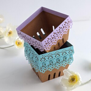 Three lace-edge paper berry baskets made from kraft cardstock, stacked and shown in lavender and soft blue colorways, styled with spring flowers on a white background, for a Lace Berry Basket SVG treat holder template for Cricut and cutting machines.