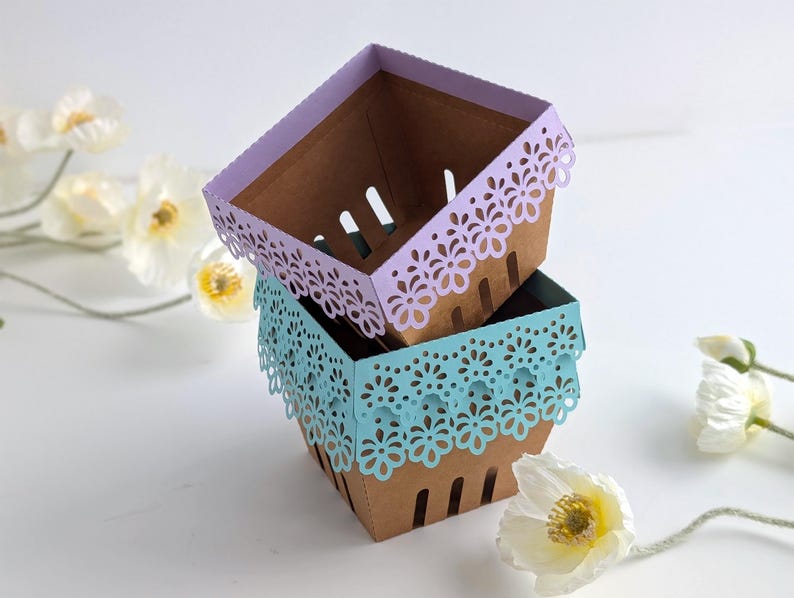 Three lace-edge paper berry baskets made from kraft cardstock, stacked and shown in lavender and soft blue colorways, styled with spring flowers on a white background, for a Lace Berry Basket SVG treat holder template for Cricut and cutting machines.