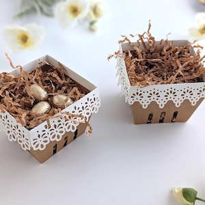 Two lace-edge paper berry baskets made from kraft cardstock, one filled with shredded paper and gold chocolate eggs and one empty, styled on a white background with soft spring flowers for a Lace Easter Basket SVG treat holder hero image.