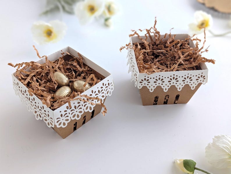 Two lace-edge paper berry baskets made from kraft cardstock, one filled with shredded paper and gold chocolate eggs and one empty, styled on a white background with soft spring flowers for a Lace Easter Basket SVG treat holder hero image.