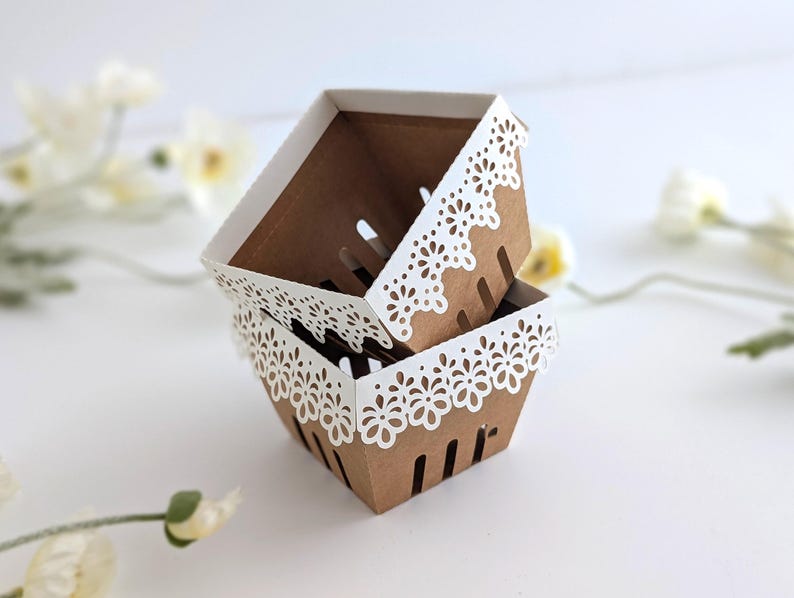 Open kraft-style berry basket with a white lace-cut paper edge, shown partially assembled on a soft neutral background with delicate flowers.
