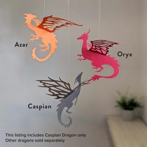 Paper dragon mobile showing three hanging dragon designs labeled Azar, Oryx, and Caspian, with Caspian dragon highlighted as the included SVG cut file and other dragons sold separately