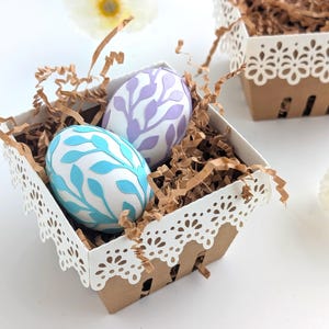 Lace-edge paper berry basket made from kraft cardstock, filled with shredded paper and two decorated Easter eggs in soft blue and lavender, styled on a white background for a Lace Easter Basket SVG treat holder template for Cricut and cutting machines.