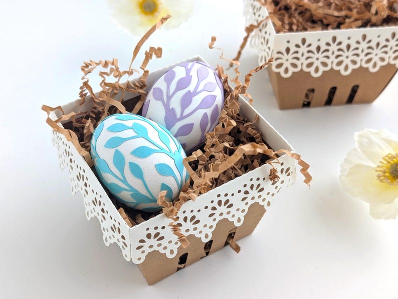 Lace-edge paper berry basket made from kraft cardstock, filled with shredded paper and two decorated Easter eggs in soft blue and lavender, styled on a white background for a Lace Easter Basket SVG treat holder template for Cricut and cutting machines.