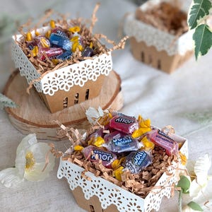 Lace Berry Basket paper treat boxes filled with candy and shredded paper filler, displayed on wood slices with flowers and greenery. Decorative lace-cut edges make them ideal for party favors, treats, and dessert table displays.