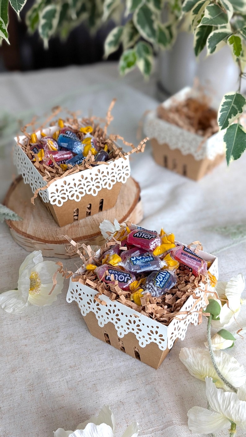 Lace Berry Basket paper treat boxes filled with candy and shredded paper filler, displayed on wood slices with flowers and greenery. Decorative lace-cut edges make them ideal for party favors, treats, and dessert table displays.