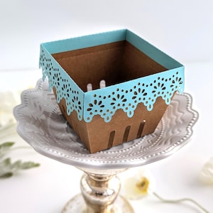 Empty lace-edge paper berry basket made from kraft cardstock with a soft blue lace rim, styled on a decorative pedestal plate with spring flowers, shown as a Lace Berry Basket SVG treat holder template for Cricut and cutting machines.