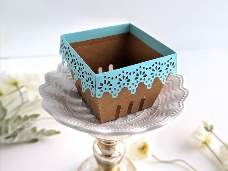 Empty lace-edge paper berry basket made from kraft cardstock with a soft blue lace rim, styled on a decorative pedestal plate with spring flowers, shown as a Lace Berry Basket SVG treat holder template for Cricut and cutting machines.