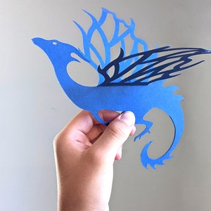 May include: A blue paper cutout of a dragon with black wings. The dragon is holding a small, white object in its mouth.