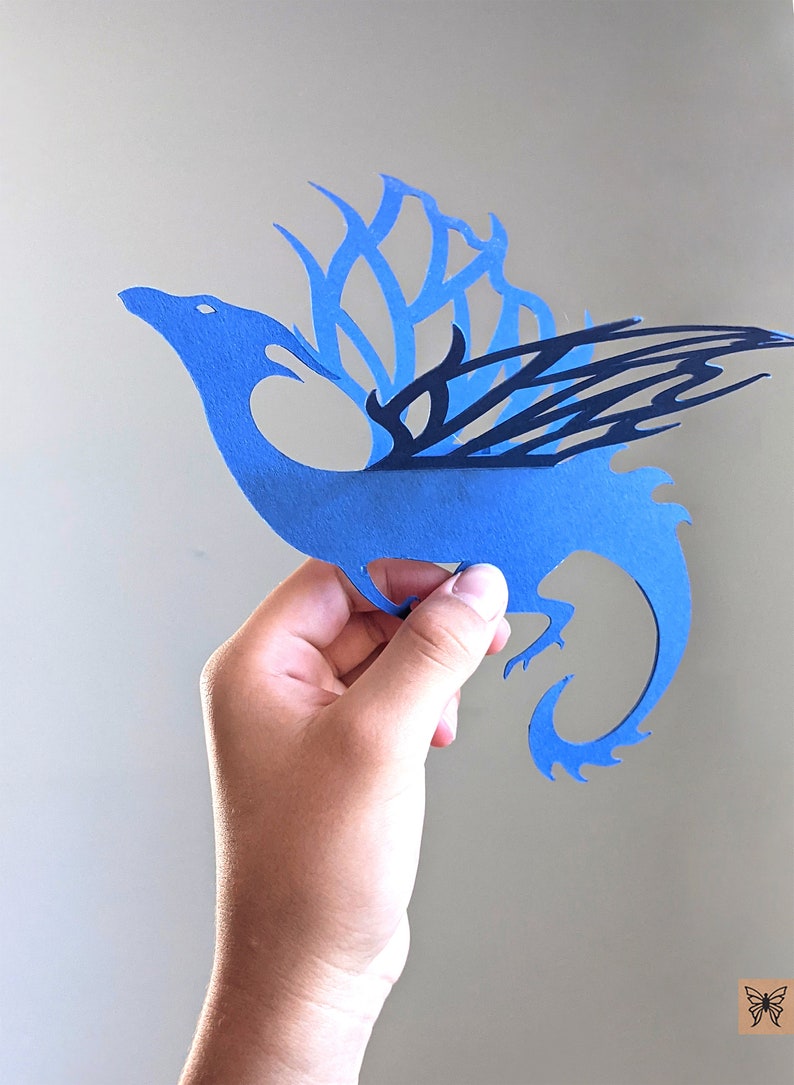 May include: A blue paper cutout of a dragon with black wings. The dragon is holding a small, white object in its mouth.