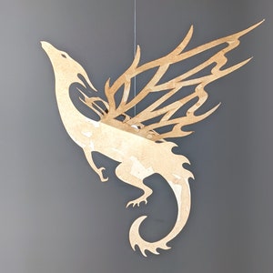 May include: A beige paper dragon mobile with intricate cut-out wings and a curled tail. The dragon is suspended from a string.