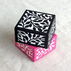 Puede incluir: Dos cajas cuadradas apiladas una encima de la otra. La caja superior es negra con un patrón floral blanco. La caja inferior es rosa con un patrón floral blanco.
