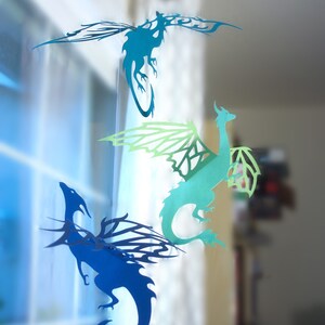 May include: Three paper dragon cutouts hanging from a window. The dragons are blue, green, and teal. The dragons are all facing the right side of the image.