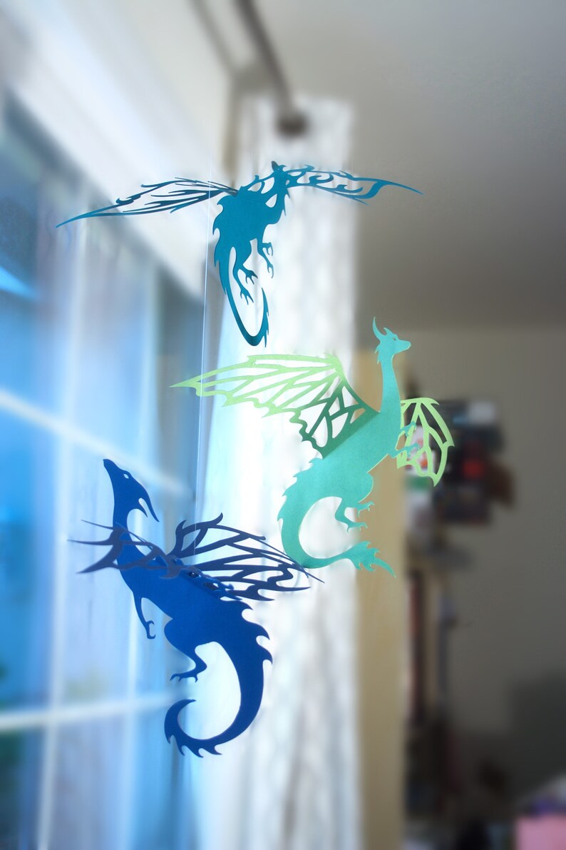 May include: Three paper dragon cutouts hanging from a window. The dragons are blue, green, and teal. The dragons are all facing the right side of the image.