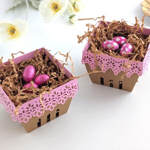 Two lace-edge paper berry baskets in pink and kraft cardstock, filled with shredded paper and pink chocolate eggs, styled on a white background with soft spring flowers for a Lace Easter Basket SVG treat holder template for Cricut and cutting machines.
