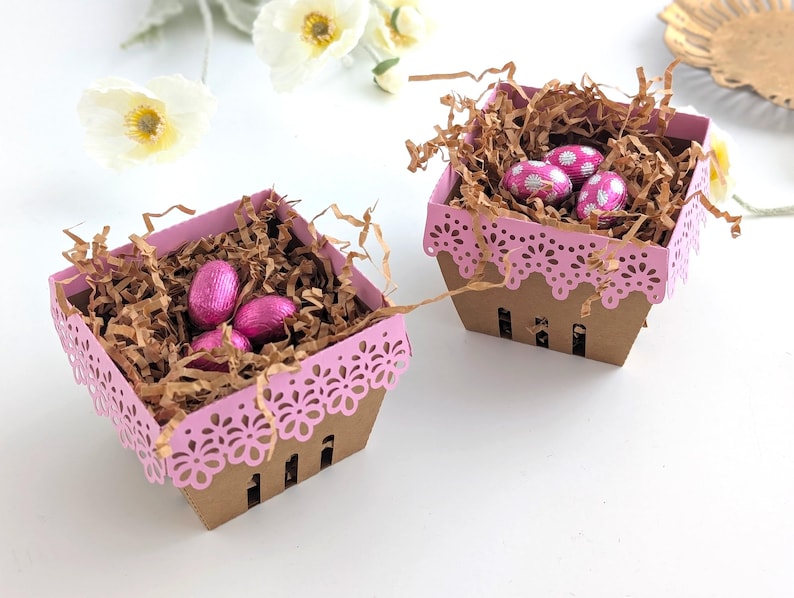 Two lace-edge paper berry baskets in pink and kraft cardstock, filled with shredded paper and pink chocolate eggs, styled on a white background with soft spring flowers for a Lace Easter Basket SVG treat holder template for Cricut and cutting machines.