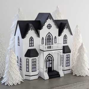 Puede incluir: Un modelo de casa en miniatura construido con papel blanco y negro, con techos a dos aguas, ventanas arqueadas y un balcón frontal. La estructura está rodeada por varios árboles de hoja perenne de papel blanco cortados con láser.