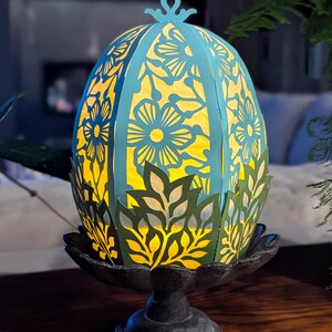 Light-up floral Easter egg basket in blue and green displayed on a pedestal stand, glowing with a flameless tealight and styled as spring table decor or Easter centerpiece.