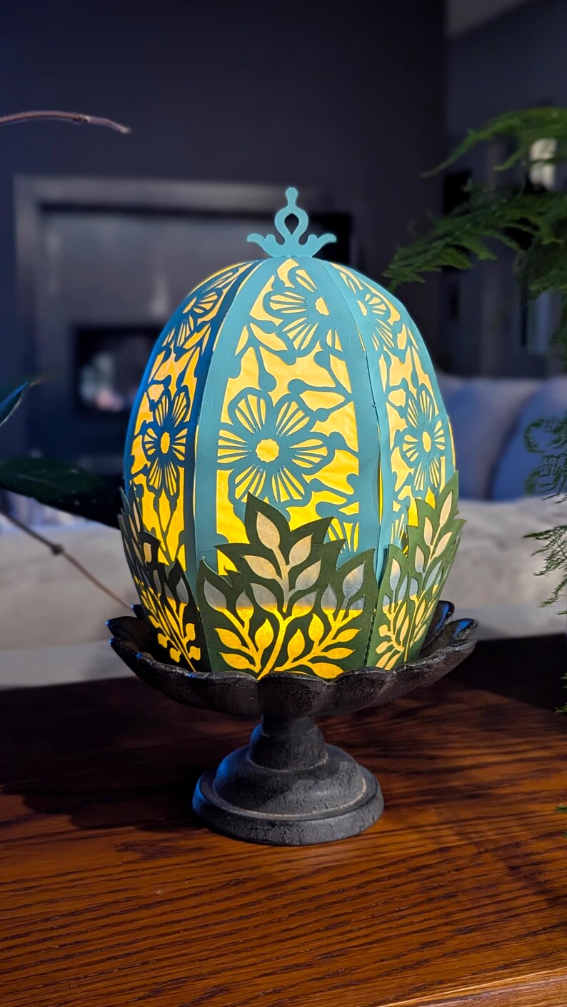 Light-up floral Easter egg basket in blue and green displayed on a pedestal stand, glowing with a flameless tealight and styled as spring table decor or Easter centerpiece.