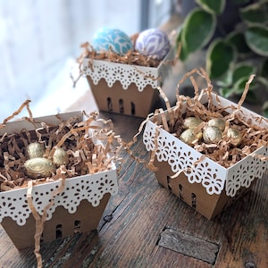 Three lace-edge paper berry baskets in white and kraft cardstock filled with shredded paper and gold and pastel decorated eggs, styled on a rustic wooden table near a window for a Lace Easter Basket SVG treat holder template for Cricut and cutting machines.