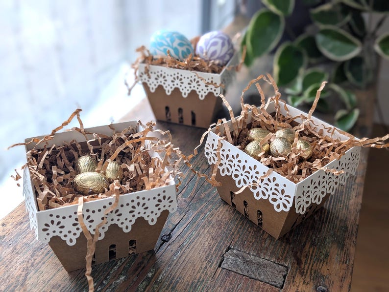 Three lace-edge paper berry baskets in white and kraft cardstock filled with shredded paper and gold and pastel decorated eggs, styled on a rustic wooden table near a window for a Lace Easter Basket SVG treat holder template for Cricut and cutting machines.