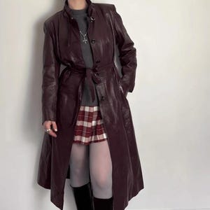 May include: A long, burgundy leather coat with a matching belt, button closure, and a high collar. The coat is paired with a plaid skirt, gray sweater, white tights, and black knee-high boots. The outfit is set against a white wall.