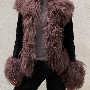May include: A black jacket with long sleeves and a fluffy, mauve-colored fur collar and cuffs. The jacket is worn over a gray top and black pants. The fur has a curly texture.