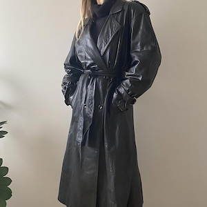 May include: A long, black leather trench coat with a belted waist and button-down front. The coat features a classic trench coat design with shoulder epaulettes and a wide lapel. The sleeves have adjustable cuffs. The coat is a vintage style.