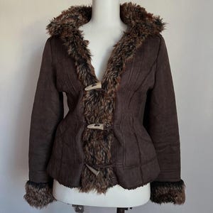 Op de afbeelding: Een bruine jas van imitatie suède met een kraag, manchetten en boorden van imitatiebont. De jas heeft houten knevelsluitingen en een getailleerde pasvorm. De bontafwerking is een mix van bruin en beige. De jas wordt getoond op een witte paspop.