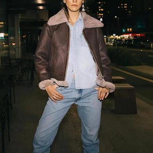 Puede incluir: Chaqueta de cuero marrón con cuello y puños de borrego. La chaqueta se lleva sobre una camisa vaquera azul claro y vaqueros azul claro. La imagen está tomada de noche con luces de la ciudad al fondo.