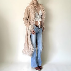 May include: A long, pale brown coat with a faux fur trim around the collar and cuffs. The coat is paired with a white top, light blue jeans, and brown boots. The coat has a suede-like texture and a flowing design.