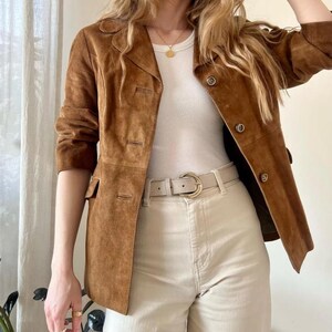 Peut inclure: Veste en daim marron avec col classique et fermeture boutonnée. La veste est associée à un haut blanc, un pantalon beige et une ceinture beige à boucle dorée. L'ensemble est complété par des bijoux dorés.