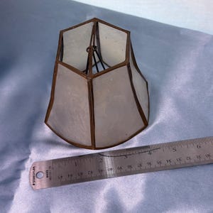May include: A hexagonal stained glass lampshade with translucent white panels and dark metal soldering. A metal ruler placed alongside shows the base of the shade measures approximately 3.5 inches in width at its widest point.