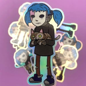 May include: Holographic sticker of a cartoon character with blue hair, a white mask, and a black outfit, holding a brown tabby cat. The sticker has a shiny, iridescent finish, with other similar stickers in the background.