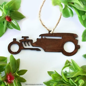 May include: A brown metal ornament in the shape of a vintage hot rod car, suspended by a natural twine string. The ornament is surrounded by green leaves and red flowers, creating a rustic and decorative appeal. The car design features a detailed engine and wheels.