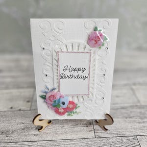 May include: A white birthday card with floral decorations and the words "Happy Birthday!". The card has a decorative frame and is supported by a wooden stand. Pink, blue, and white flowers are arranged on the card.