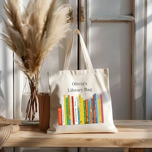 May include: A cream-colored tote bag with the text "Olivia's Library Bag" above a colorful graphic of stacked books. The bag has two handles and is sitting on a wooden surface.