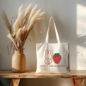 May include: Cream-colored tote bag featuring a red cartoon bunny and strawberry design with the text "MIFFY STRAWBERRY". The bag has long handles and is made of a sturdy fabric. A vase with dried ornamental grass sits to the left.