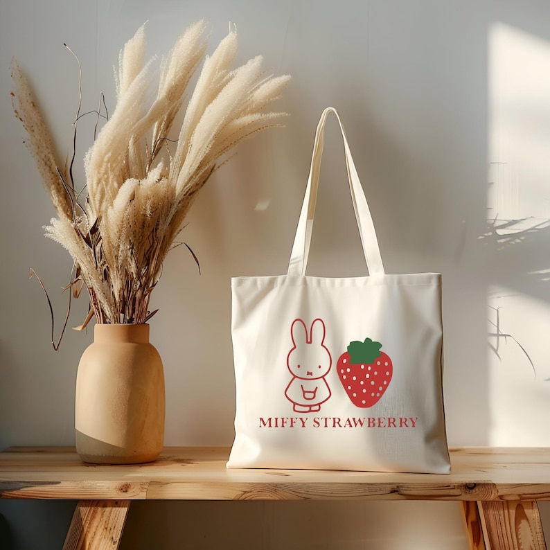 May include: Cream-colored tote bag featuring a red cartoon bunny and strawberry design with the text "MIFFY STRAWBERRY". The bag has long handles and is made of a sturdy fabric. A vase with dried ornamental grass sits to the left.