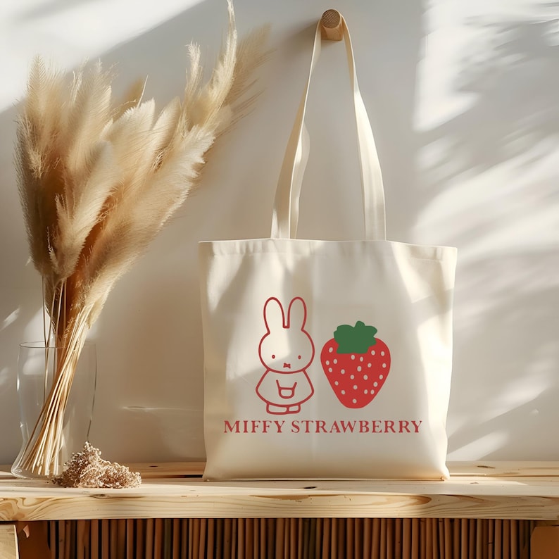 May include: Cream-colored tote bag with a red cartoon bunny and strawberry graphic. The text "MIFFY STRAWBERRY" is printed below the images. The bag hangs from a wooden peg on a white wall, with dried pampas grass in a glass vase to the left.