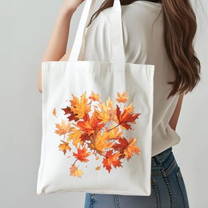 以下が含まれることがあります： オレンジ、赤、黄色で描かれた、秋の紅葉の鮮やかなイラストが特徴の白いトートバッグ。長い持ち手があり、丈夫な生地でできており、食料品やその他のアイテムの持ち運びに最適です。