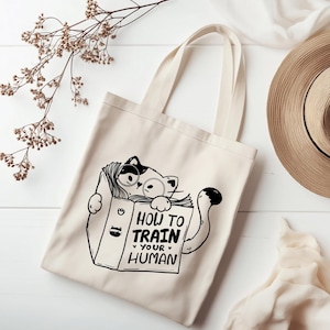 May include: Cream-colored tote bag featuring a black and white illustration of a cat wearing glasses and reading a book titled "HOW TO TRAIN YOUR HUMAN." The cat's tail is curled. The bag has two handles.