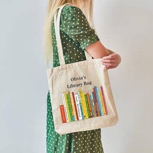 May include: Beige canvas tote bag with the text "Olivia's Library Bag" and a colorful graphic of stacked books. The bag has two handles and is being held by a person wearing a green polka dot dress.