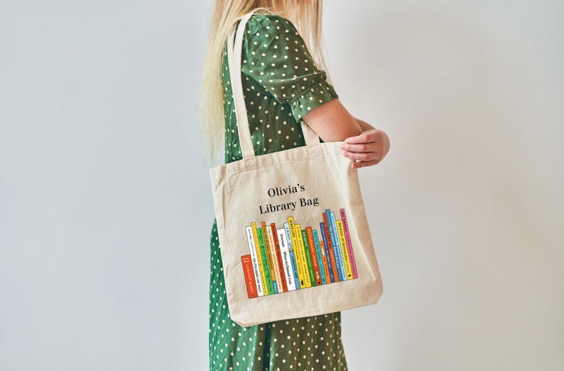 May include: Beige canvas tote bag with the text "Olivia's Library Bag" and a colorful graphic of stacked books. The bag has two handles and is being held by a person wearing a green polka dot dress.