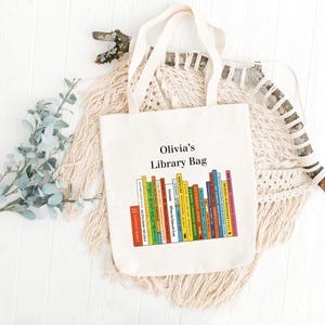 May include: A cream-colored tote bag with the text "Olivia's Library Bag" above a colorful illustration of stacked books. The bag has two handles and is made of a canvas-like material.