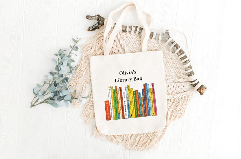 May include: A cream-colored tote bag with the text "Olivia's Library Bag" above a colorful illustration of stacked books. The bag has two handles and is made of a canvas-like material.