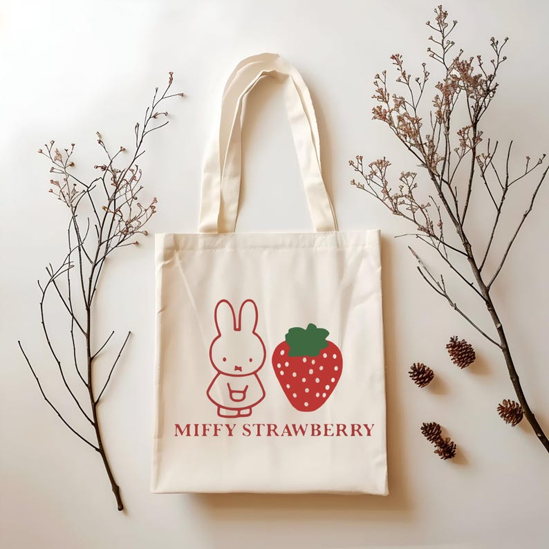 May include: Cream-colored tote bag featuring a red cartoon bunny and a red strawberry with green leaves. The words "MIFFY STRAWBERRY" are printed below the illustrations. The bag has two cream-colored handles.