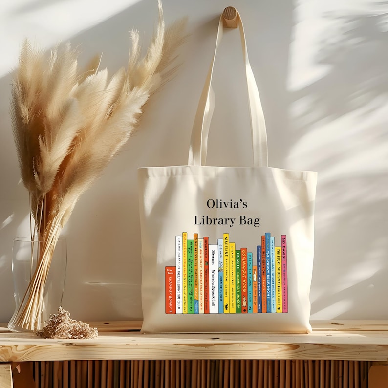 May include: Cream-colored tote bag with the text "Olivia's Library Bag" above a colorful graphic of stacked books. The bag has long handles and is hanging on a wooden peg. A vase of dried flowers is to the left.