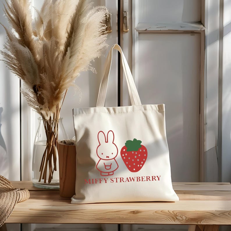 May include: Cream-colored tote bag featuring a red outline illustration of a bunny and a strawberry. The words "MIFFY STRAWBERRY" are printed in red below the images. The bag has two handles and is made of a sturdy fabric.