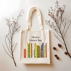 May include: Cream-colored tote bag with the text "Olivia's Library Bag" above a row of colorful book spines. The bag has two handles and is decorated with sprigs and pinecones.